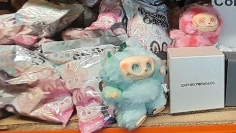 A blue fake Labubu doll, pink doll in backround, beside numerous plastic bags and a silver box with Armani logo on it