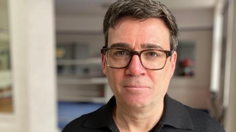 Andy Burnham, with short black hair, wears glasses and a black T-shirt and jacket with a yellow bee motif badge on it