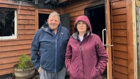 A man and woman in front of a wooden garden shed. It is raining and they both have their hands in their pockets. The woman is wearing a purple rain coat and the man is in a blue coat. 