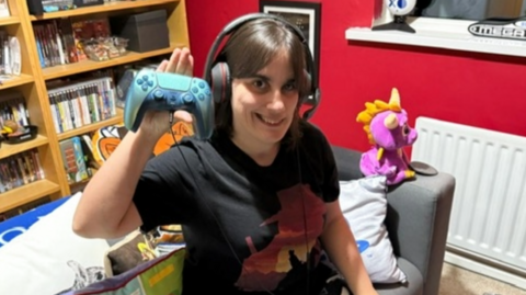 Louise Meeke has long brown hair and is wearing a black T-shirt with a cowboy motif. She is wearing a large black headset and holding a blue PS5 controller in her right hand. A Spyro the Dragon toy can be seen on the grey sofa beside her. Several games are visible in a library bookshelf nearby.