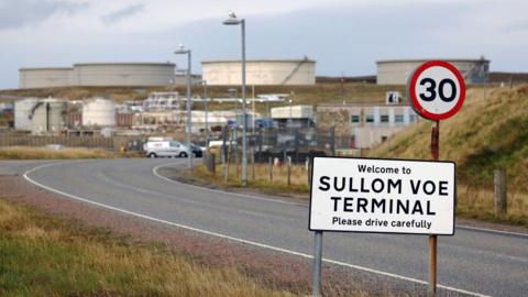 A 30mph sign which says 'Welcome to SULLOM VOE TERMINAL - Please drive carefully', with oil and gas infrastructure in the background after a winding road.