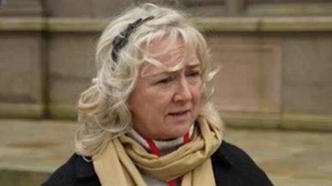 Deputy Karen Wilson is wearing a black jacket and a beige scarf. She has blonde hair and is stood outside a stone building