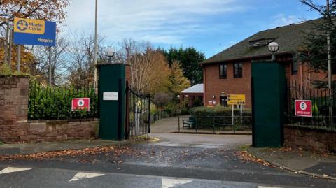 Marie Curie hospice is a two-storey brick building set behind a sandstone wall and large gates. A blue sign and yellow sign has the writing Marie Curie Hospice. There are two red traffic warning signs which say 5 speed ramp.