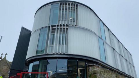 The front of a white curved three-storey building which has two large windows at the front. On the right-side of the building is more windows. On the bottom floor is large glass windows with a red bar in front. On the left is the top of a chapel. 