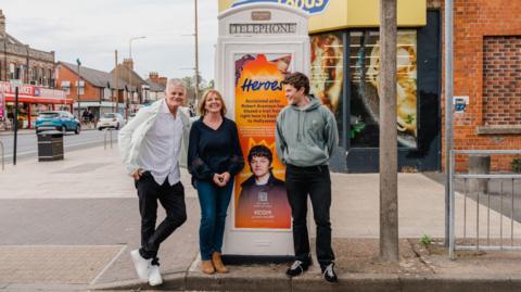 Three people - a man and his parents - stand either side of a cream mid-20th Century telephone box in a broad high street. The phone box is decorated with a colourful poster featuring a picture of the actor and the words: "Hometown heroes - acclaimed actor Rob Aramayo has blazed a trail from right here in East Hull to Hollywood". The man is wearing a green hoodie, black jeans and black trainers. He is smiling as he looks at his parents. His father is leaning against the phone box and wearing a cream jacket, white shirt, black jeans and white trainers. His mother, who has strawberry blonde hair, is wearing a blue top, matching jeans and brown boots.