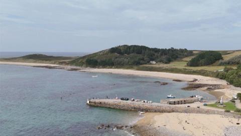 Aerial view of a quiet coastal bay with clear blue-green water, sandy and rocky beaches, and a small stone pier extending into the sea. A cluster of white buildings sits among lush gardens and trees on the shoreline, while rolling green hills and wooded areas rise in the background beneath a lightly clouded sky.