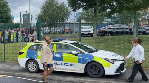 Outside shot of the school where it took place. A parked police car is seen , in front of the school gates. Two people walk past.