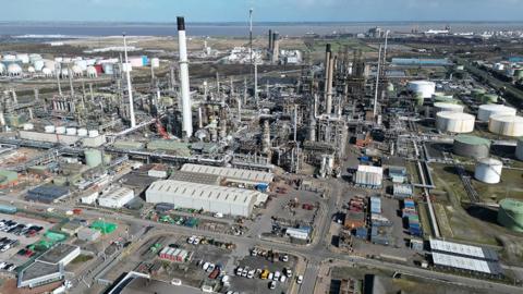 Aerial view of the Humber Refinery it has a number of white tanks and machinery with pipes and and chimneys. A port with ships docked can be seen in the background