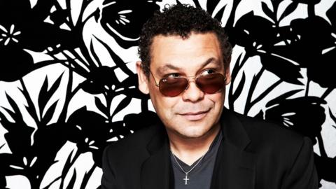 Craig Charles looks to the side, he is wearing sunglasses and smiling subtly. He is sitting against a floral, black and white background. © BBC/Dean Chalkley