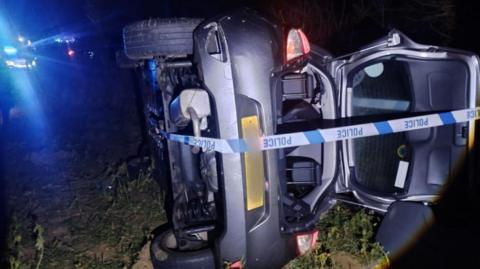 A car on its side in a field in the dark wrapped in police tape