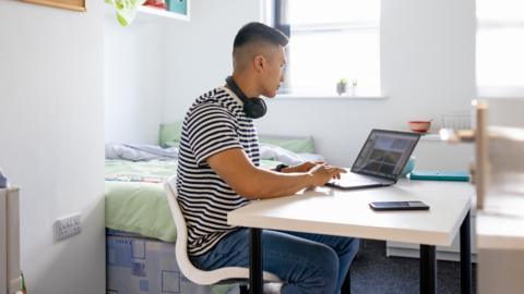 A man in a black and white striped t-shirt and jeans sits at a white desk using a laptop