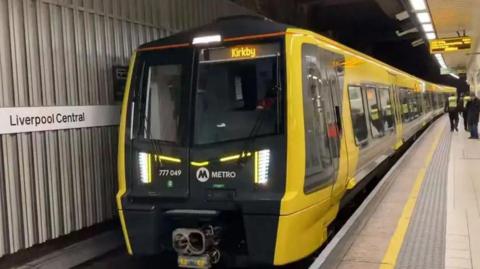 A yellow and black Merseyrail train at a platform at Liverpool Central station. The destination Kirkby is displayed in yellow at the front of the train which carries the Metro logo.