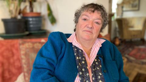 A woman with short, curly grey hair and wearing a pink and white striped shirt and a blue cardigan sits in a living room.