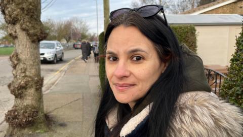 Yola Caprin is standing in front of a gate which is on the right. She has long black hair and black sunglasses on her head. She is wearing a khaki hoodie and fur coat. 