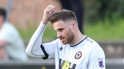 David Goodwillie playing for Glasgow United FC. He is wearing a white top with the team badge on it. His right arm is raised and his behind his head.