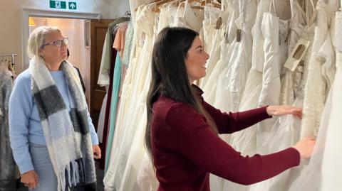 A woman with long brown hair sorts through hanging wedding dresses while a woman with a blue jumper and scarf watches behind her