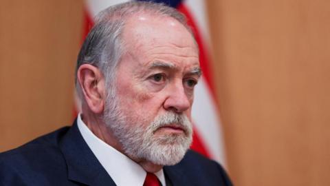 Mike Huckabee, who has grey hair and a short grey beard and moustache, looks on during an interview at his office at the US embassy in Jerusalem. An American flag is behind him.