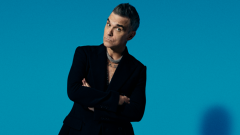 Robbie Williams is standing in front of a blue background. He has crossed his arms and is raising his eyebrows at the camera. He is wearing a black jacket.