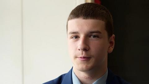 A head and shoulders image of a young man. He is wearing a navy blazer over a smart shirt. He is looking into the camera.