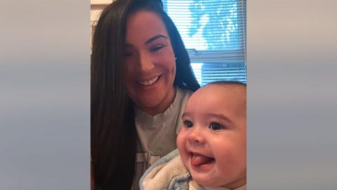 Sasha Field with long black hair, who is smiling as she is holding a baby who is also smiling with his tongue sticking out.