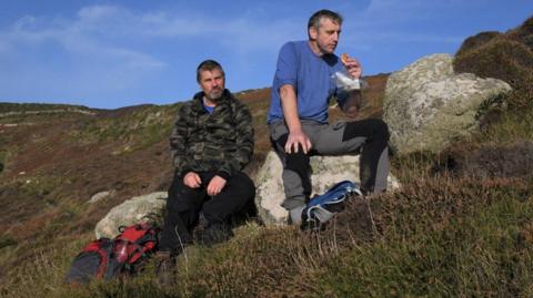 Two men sitting on a hill in Cornwall. One is waring a military-pattern jacket, black trousers and a red backpack. The other is wearing a blue t-shirt and dark grey trousers. 