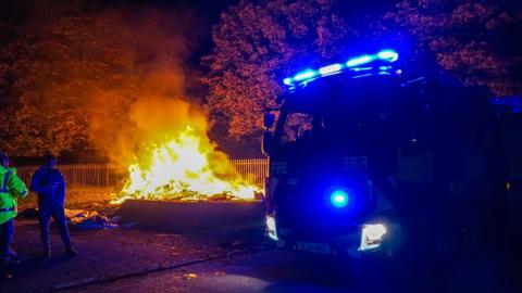 Fire engine next to bonfire in the dark with two people standing nearby. One of the people is wearing a high-vis jacket. 