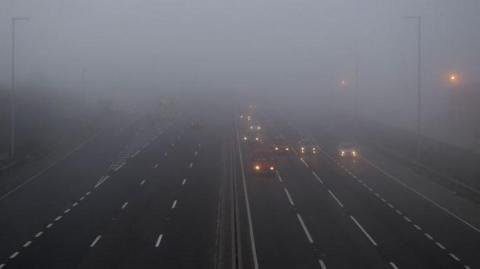 Cars driving through fog on a motorway. There are three lanes of traffic on either side and the headlights of the vehicles are fuzzy and some are barely visible due to the foggy conditions.
