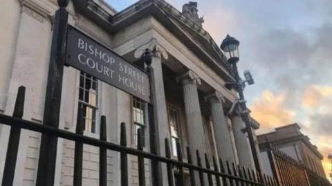 The outside of Bishop Street Court House in Londonderry. A street lamp and black gate is just outside a pillared building.