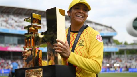Trent Rockets batter Sophia Dunkley smiles as she hold up the 2026 Hundred trophy