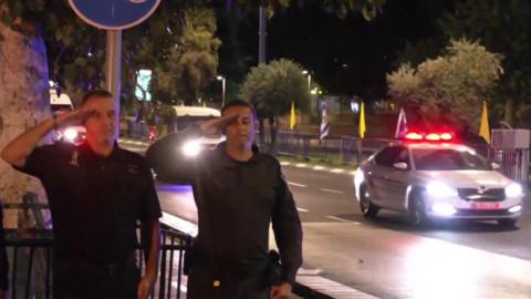 Two men salute as a police convoy with flashing red lights can be seen approaching behind them.