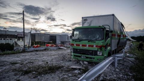 A damaged truck on a debris-covered road following an explosion at the Aeon Mall which is in the background