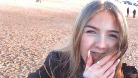 Lucy Hill on a beach covering half of her mouth with her hand, it has a ring on it and she is smiling 