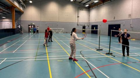 People playing badminton in a large hall inside a leisure centre. The floor is blue with multi-coloured lines.