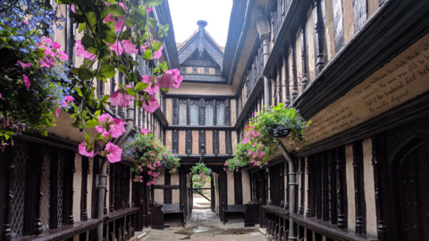 A courtyard of a Tudor building with pink hibiscus flowers in hanging baskets. There is an open doorway ahead, with rooms above it and the pitched roof of the building can also be seen. 