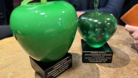 Two ornament green apples, one larger than the other, being used as trophies, so sitting on engraved plinths.