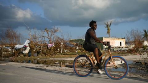 A woman rides a bicycle past damage in a destroyed neighborhood in the wake of Hurricane Dorian in Marsh Harbour