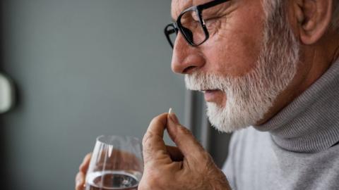 A man with a white beard holds a glass of water while taking a pill