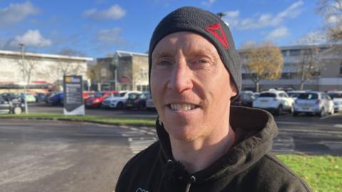 Steve Bunn looks into the camera, wearing a woolly hat and hoodie in front of the leisure centre car park.