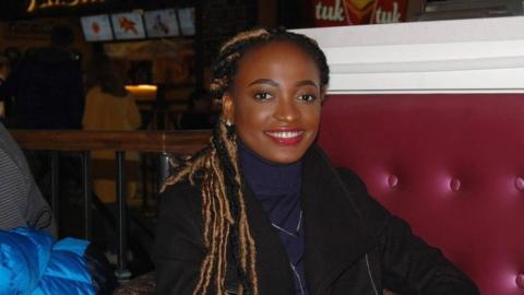 A woman with long hair styled in locs sits in a cafe or diner with red cushioned walls and signs advertising various food visible in the background. She's smiling and wearing a navy blue polo-necked top underneath a black jacket. She wears red lipstick and small, sparkly silver stud earrings.