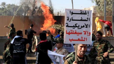 A man holds up a sign saying "Close the American embassy or people will close it" as protesters set fire to the wall of the US embassy compound in Baghdad, Iraq (31 December 2019)