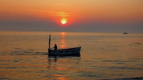 The sky is layers of red and orange with a burning yellow setting sun in the centre. It’s all reflected in the sea below with a person on a boat in the middle of the image.