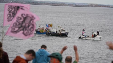 People on shore wave flags at boats on water