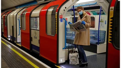 A woman wearing PPE (personal protective equipment), including a face mask as a precautionary measure against COVID-19, reads a newspaper as she stands aboard a London Underground Tube train, i