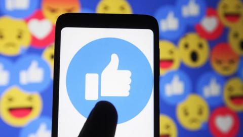 In this photo illustration a thumb up sign emoji and smiley face icons of Facebook are seen displayed on a mobile phone and a pc screen.