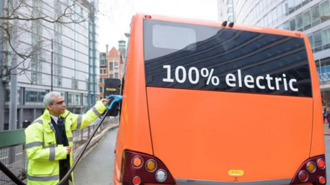 The back of an orange bus. In the top window it reads "100% electric". A man wearing a high vis jacket.