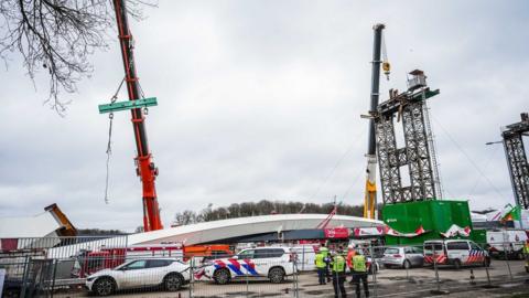 Emergency services on the scene of the bridge collapse at a construction site. Cranes and a partially constructed bridge are visible behind.