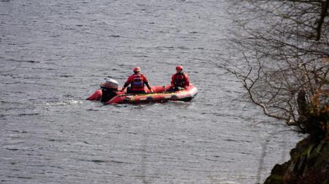 Two volunteers from Patterdale Mountain Rescue Team on a small motor boat. They are searching Ullswater for the missing swimmer. They are wearing red protective clothing and helmets.