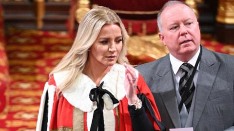A woman with long blonde hair is dressed in a fur trimmed red and gold gown and her middle aged husband is in a grey three-piece suit. They are standing inside a regal looking room with red and gold carpets