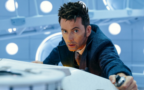 A man with spiky brown hair wearing a blue suit, white shirt and purple tie. He is leaning over a white piece of machinery, holding a piece of metal. Behind him are bright white lights.