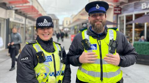 PC Jill Robertson and PCSO Dan Snell on patrol in Workington town centre. They are both in uniform, with hats and high-viz vests. They are standing next to each other and smiling at the camera. PC Robertson's hair is tied back. PCSO Snell has a beard and septum piercing.